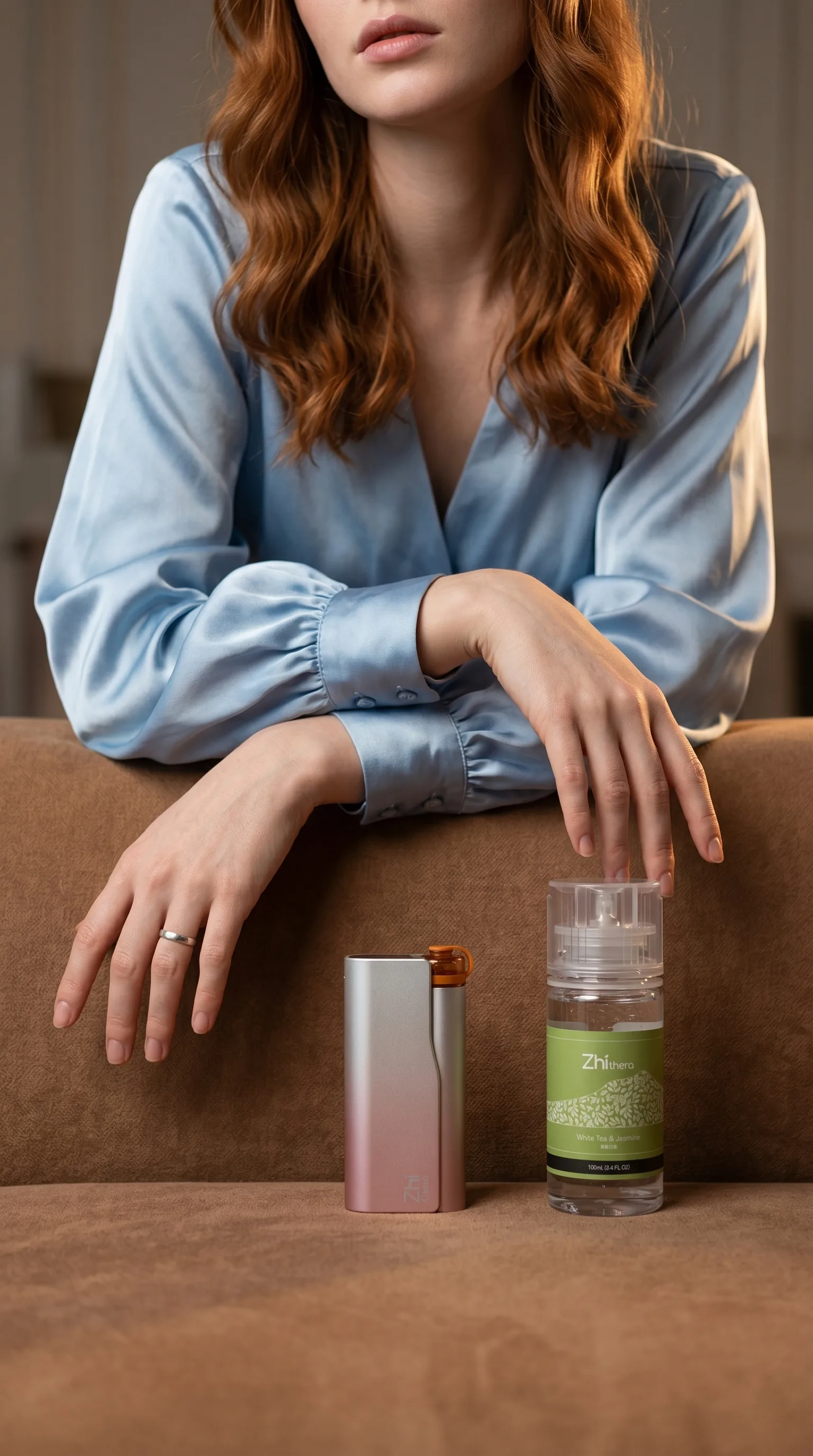 A woman with wavy red hair wearing a light blue silk blouse rests her arms on a brown sofa while reaching toward a silver…