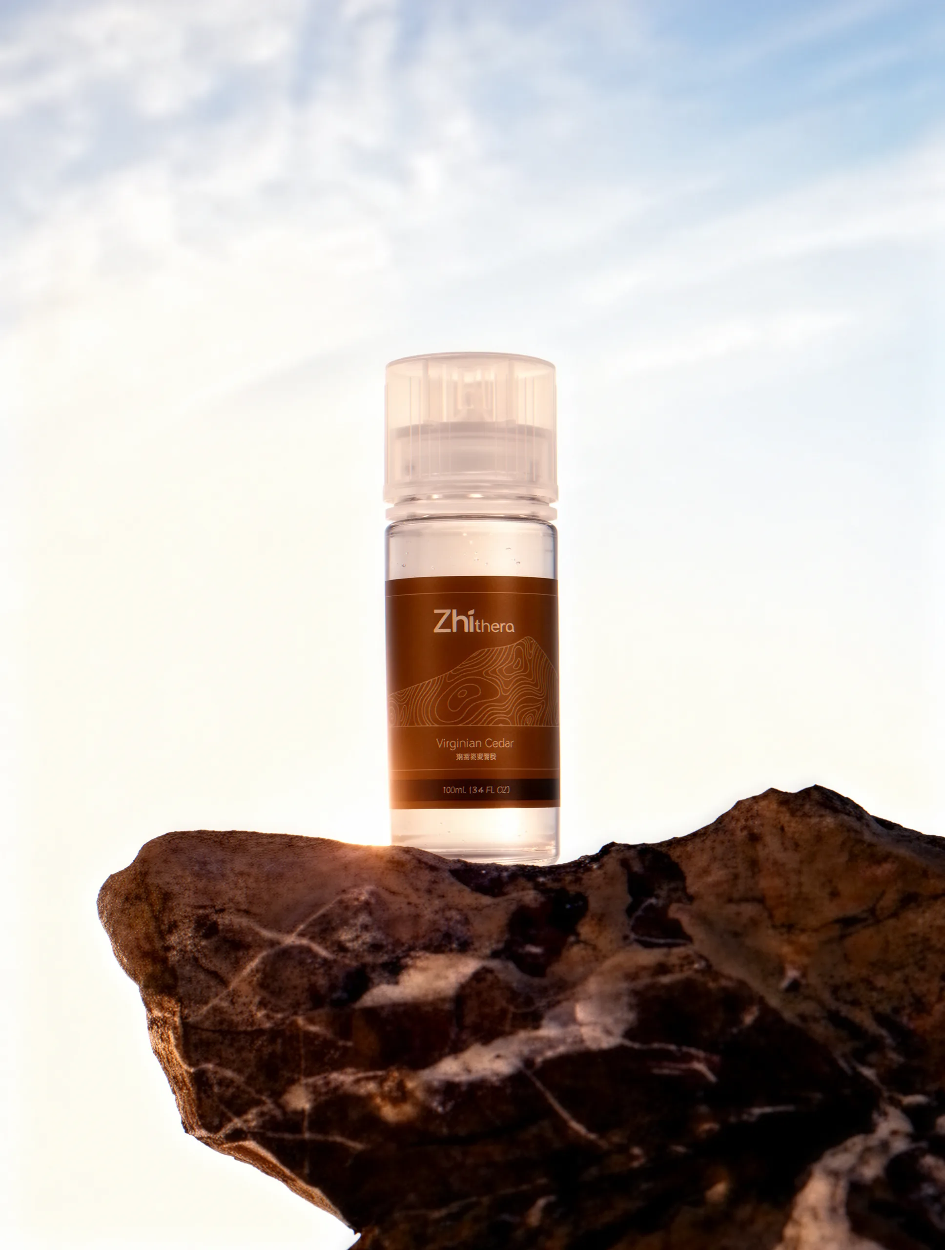 A clear glass bottle with an amber label sits atop a rugged, dark brown rock against a bright, sunlit sky, evoking a sense…